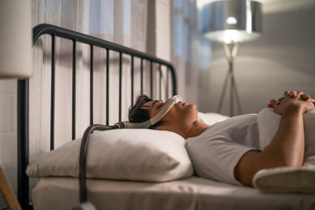 Homem deitado utilizando CPAP. Refletindo sobre: A apneia obstrutiva do sono é frequentemente desencadeada por uma combinação de fatores, sendo a obesidade um dos principais contribuintes.